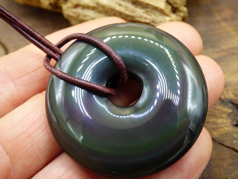 K&ouml;nnte beinhalten: Ein dunkler gr&uuml;ner Obsidian-Donut-Anh&auml;nger mit einem Lederband. Der Anh&auml;nger hat einen hohen Glanz und einen irisierenden Schimmer.