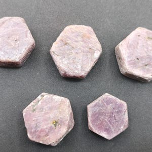 May include: Five loose, hexagonal-shaped gemstones with a pale, dusty pink and lavender hue. Each stone features a flat, matte surface with visible crystalline textures and natural inclusions, arranged in a scattered formation.