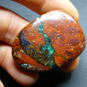 May include: A polished, rounded stone with a rich, earthy color palette. The stone features a rusty-red base, accented by streaks of deep blue, emerald green, and black. Small, light-colored inclusions dot the surface, adding texture.