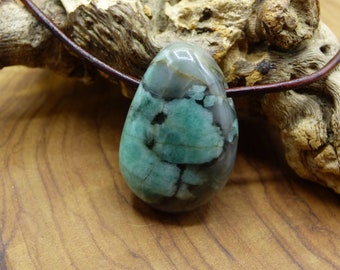 May include: A teardrop-shaped pendant with a green and gray marbled pattern, strung on a brown leather cord. The gemstone is set against a backdrop of weathered wood.