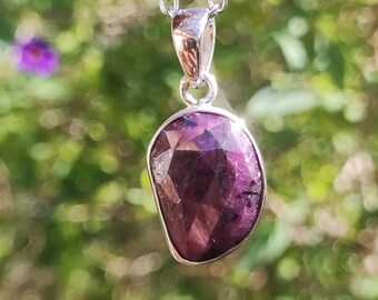 May include: A silver pendant necklace featuring a faceted, oval-shaped, deep purple gemstone. The pendant hangs from a delicate silver chain. The background is a soft focus of green foliage and purple flowers.