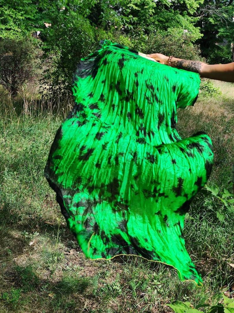 May include: A vibrant green and black tie-dye fabric fan, held outdoors. The fan's fabric is a bright, eye-catching green with irregular black splotches. The fan is open, displaying its full, flowing form against a backdrop of green foliage.