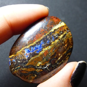 May include: A polished, oval-shaped gemstone with a striking pattern of colors. The stone features bands of brown, red, yellow, and blue hues. The gemstone is held between two fingers, showcasing its natural beauty and unique color variations.