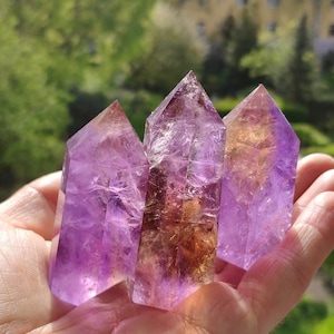 Pode incluir: Três pontas de cristal de ametista com um tom marrom dourado. Os cristais estão dispostos em forma triangular e são mantidos em uma mão.