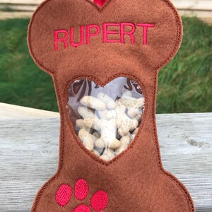 May include: A brown felt dog bone-shaped treat holder with a clear heart-shaped window. The holder has the name "RUPERT" embroidered on the top and a red paw print on the bottom.