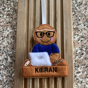 Puede incluir: Un adorno de pan de jengibre en forma de persona con gafas, que lleva una camisa azul y está sentado en un escritorio con un portátil. El adorno tiene el nombre "Kieran" en el escritorio.