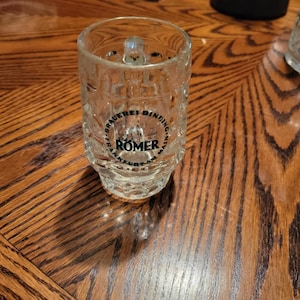 May include: A clear glass beer mug with a handle, featuring the text "Brauerei Binding Frankfurt a.M. Romer". The mug has a textured design and is sitting on a wooden table.