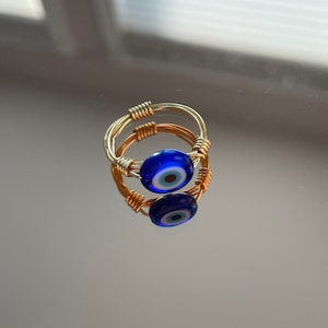 May include: A handmade ring featuring a vibrant blue evil eye bead, set in a gold-toned wire frame. The ring's design includes intricate wire wrapping and a reflective surface, showcasing the ring's details.