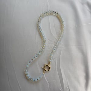 May include: A delicate necklace featuring small, round, opalescent beads strung together. The necklace has a gold-colored clasp and small gold-colored beads interspersed throughout the strand. The beads are a soft, translucent white color.