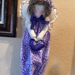 May include: A purple and white fabric bag with a floral pattern. The bag is shaped like a dress and has a white fabric angel wing on each side. The angel has a grey hair wig and a purple bow. The bag is holding a small purple heart-shaped bag.