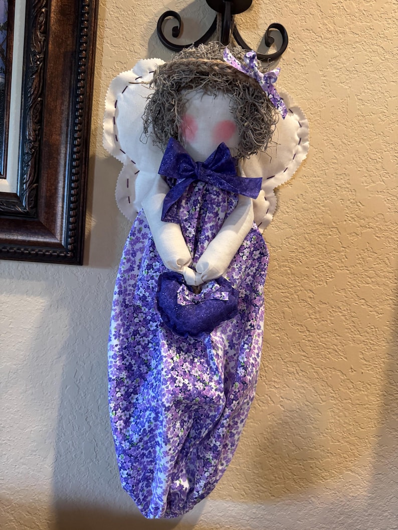 May include: A purple and white fabric bag with a floral pattern. The bag is shaped like a dress and has a white fabric angel wing on each side. The angel has a grey hair wig and a purple bow. The bag is holding a small purple heart-shaped bag.