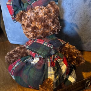 May include: A brown teddy bear dressed in a plaid outfit. The dress and bow are made from a patchwork of red, green, and black plaid fabric. The bear has brown fur and a small, fluffy tail, creating a classic, cozy aesthetic.