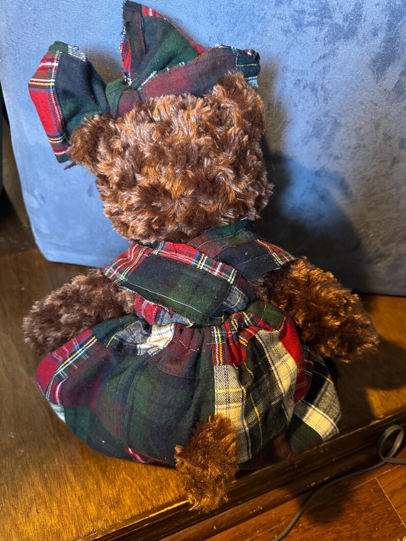 May include: A brown teddy bear dressed in a plaid outfit. The dress and bow are made from a patchwork of red, green, and black plaid fabric. The bear has brown fur and a small, fluffy tail, creating a classic, cozy aesthetic.