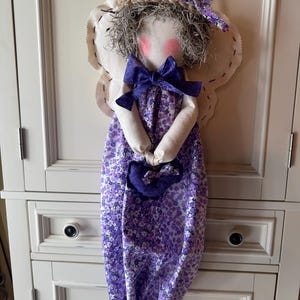 May include: A reusable grocery bag with a handmade doll attached. The bag is made of purple and white floral fabric. The doll has a white face, pink cheeks, brown hair, and a purple bow. The doll is wearing a white dress with a purple floral print.