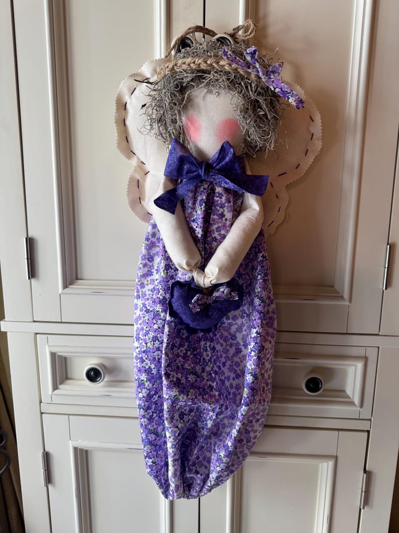 May include: A reusable grocery bag with a handmade doll attached. The bag is made of purple and white floral fabric. The doll has a white face, pink cheeks, brown hair, and a purple bow. The doll is wearing a white dress with a purple floral print.