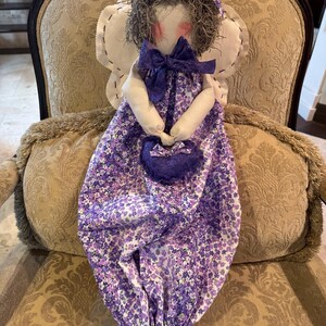 May include: A handmade fabric angel doll with a purple floral dress, a purple bow, and a purple purse. The doll is sitting on a chair with a floral pattern.