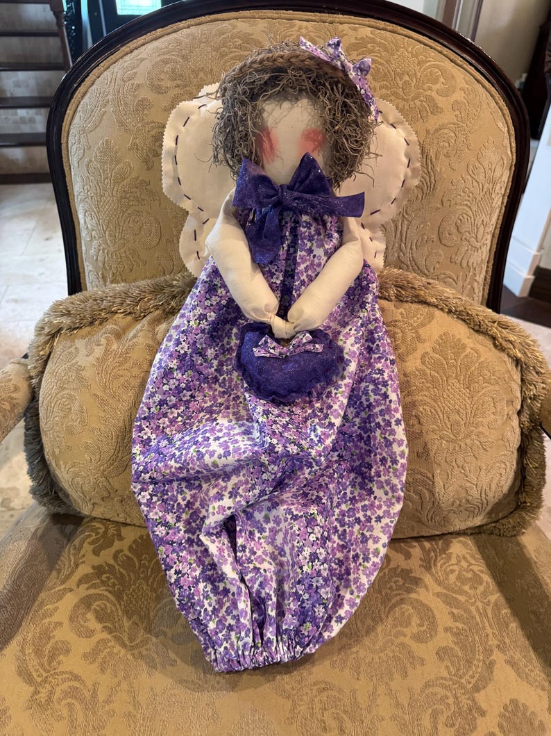 May include: A handmade fabric angel doll with a purple floral dress, a purple bow, and a purple purse. The doll is sitting on a chair with a floral pattern.
