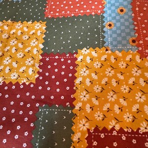Op de afbeelding: Een stof met een patchworkontwerp met gekartelde randen. Het patroon bestaat uit rechthoekige secties in donkergroen, mosterdgeel, dieprood en lichtblauw, versierd met kleine witte bloemenprints en zonnebloemmotieven.