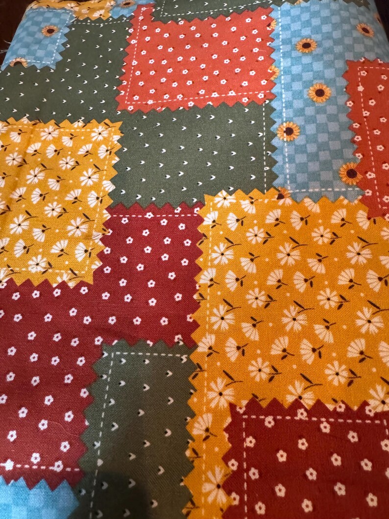 Op de afbeelding: Een stof met een patchworkontwerp met gekartelde randen. Het patroon bestaat uit rechthoekige secties in donkergroen, mosterdgeel, dieprood en lichtblauw, versierd met kleine witte bloemenprints en zonnebloemmotieven.