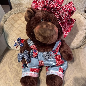 Op de afbeelding: Een bruine pluche teddybeer met een grote rode strik met bandana-print op het hoofd en een patchwork tuinbroek met rode, witte en blauwe patronen. De beer houdt een klein blauw-wit windmolentje met sterrenpatroon in de rechterpoot.