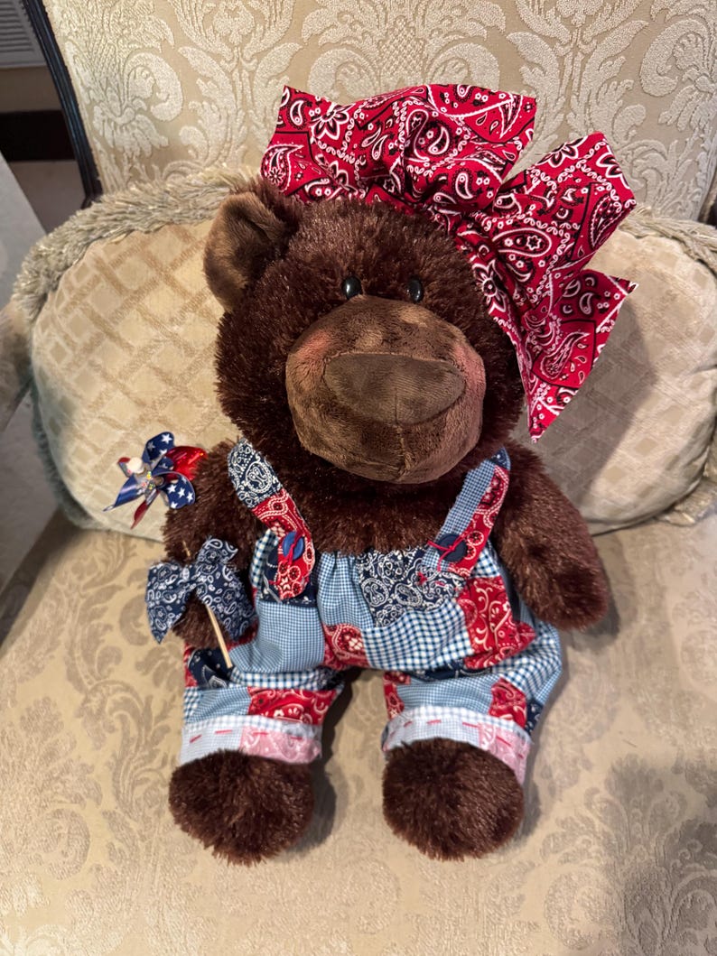 Op de afbeelding: Een bruine pluche teddybeer met een grote rode strik met bandana-print op het hoofd en een patchwork tuinbroek met rode, witte en blauwe patronen. De beer houdt een klein blauw-wit windmolentje met sterrenpatroon in de rechterpoot.