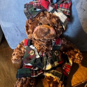 May include: A brown teddy bear with a large plaid bow and matching plaid overalls. The bear has a pink snout and button eyes. The plaid pattern includes red, green, and white. The bear is sitting on a wooden surface.