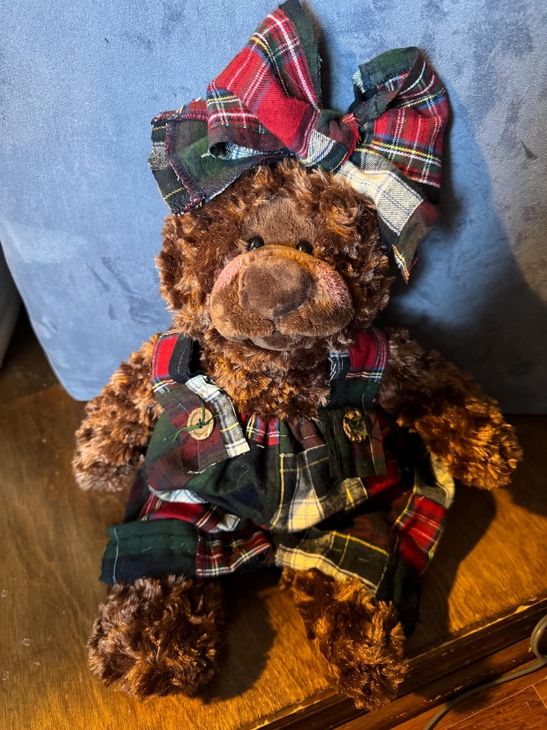May include: A brown teddy bear wearing a large plaid bow and matching plaid overalls. The bear has a pink snout and button eyes. The plaid pattern includes red, green, and white. The bear is on a wooden surface.