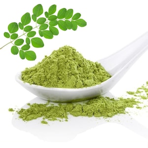 May include: A white spoon filled with green moringa powder, with a sprig of moringa leaves in the background.