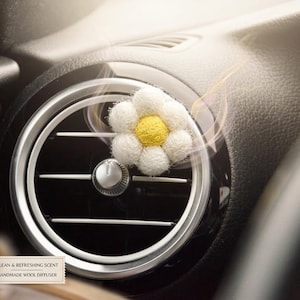 May include: A car air vent with a daisy-shaped handmade wool diffuser. The daisy has white petals and a yellow center. A small glass bottle of essential oil with a wooden cap sits below the vent. The text on the label reads "Lavender Fields Essential Oil".