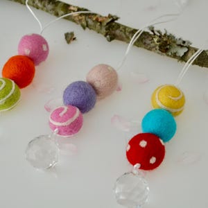 May include: Colorful felt ball garlands with crystal accents. The garlands feature various colors, including pink, orange, green, purple, and red, with white accents. The garlands are strung on white string and are attached to a natural branch.