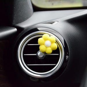 May include: A yellow and white flower-shaped car air vent clip. The flower is made of soft, fuzzy material and is attached to a silver and black car vent. The car's interior is black.