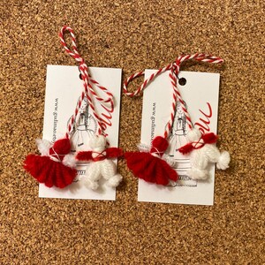 May include: Two handmade red and white yarn dolls with matching tags. Each tag features a drawing of a woman in traditional clothing and the text "me". The dolls are attached to the tags with red and white twine.