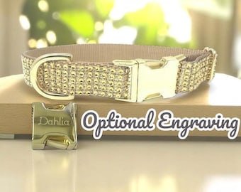 May include: A tan dog collar with gold-colored hardware and a buckle. The collar is embellished with small, gold-colored rhinestones. A separate gold-colored buckle is shown with the name "Dahlia" engraved on it. The text "Optional Engraving" is also visible.