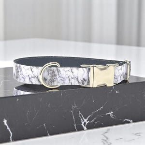 May include: A dog collar with a marble pattern in white and gray, featuring a silver-colored D-ring and a gold-colored metal buckle. The collar has a dark gray interior and is displayed on a black marble surface.