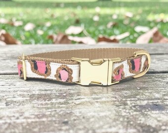 May include: A tan dog collar with a gold-colored buckle. The collar features a white band with pink and brown leopard print accents. The collar is made of durable material and is designed for pets.