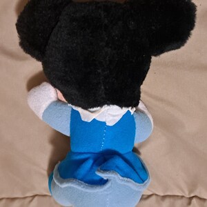 May include: A plush toy of a cartoon character with black fur, large ears, and a blue dress. The toy is lying on its side with its head resting on its arm.