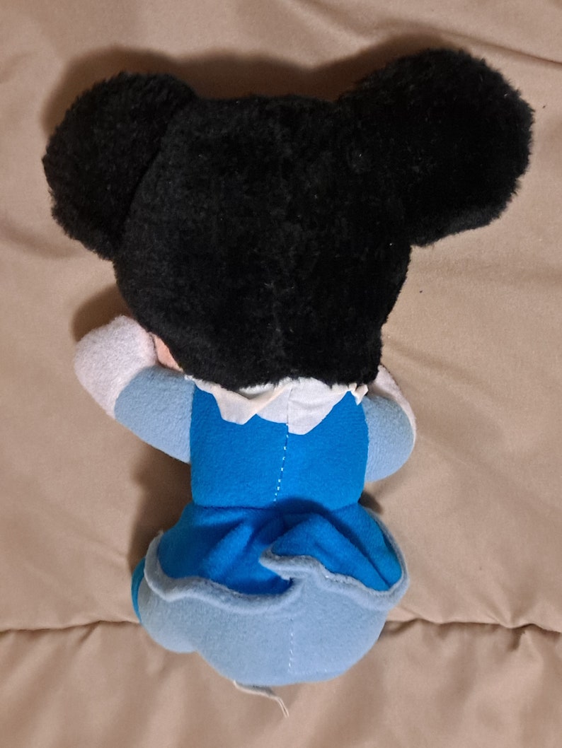 May include: A plush toy of a cartoon character with black fur, large ears, and a blue dress. The toy is lying on its side with its head resting on its arm.