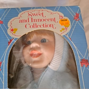 May include: A blue and white porcelain doll with blonde hair and blue eyes. The doll is wearing a blue hooded jacket with a white trim. The doll is packaged in a blue box with the words "Sweet and Innocent Collection" printed on it. The price tag on the box reads "$19.99".
