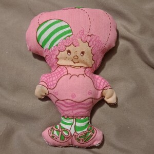 May include: A pink and green plush doll with a green and white striped hat. The doll has a pink dress with white polka dots and green and white striped socks.