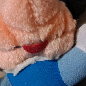 May include: A plush toy with a pink face, black ears, and a red mouth. The toy is wearing a blue and white shirt.
