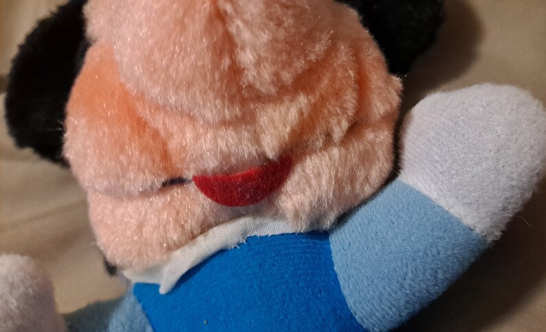 May include: A plush toy with a pink face, black ears, and a red mouth. The toy is wearing a blue and white shirt.