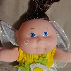 May include: A Cabbage Patch Kid doll dressed as a fairy with a yellow dress, green leaves, and a white flower. The doll has brown hair, blue eyes, and a daisy painted on its cheek.