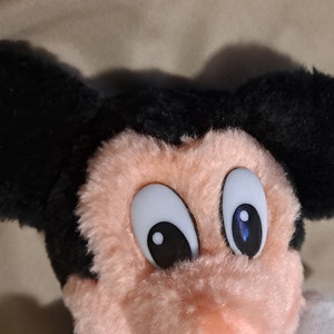 May include: A plush Mickey Mouse toy with black ears and a pink face. The toy has large white eyes with black pupils and blue accents.
