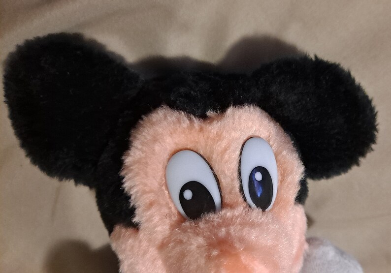 May include: A plush Mickey Mouse toy with black ears and a pink face. The toy has large white eyes with black pupils and blue accents.