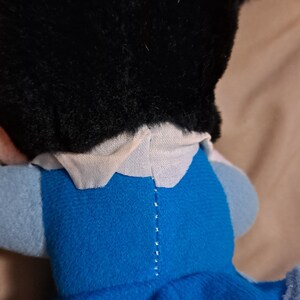 May include: A plush toy with black hair, a white collar, and a blue dress. The toy is lying on its back with its head turned to the side.