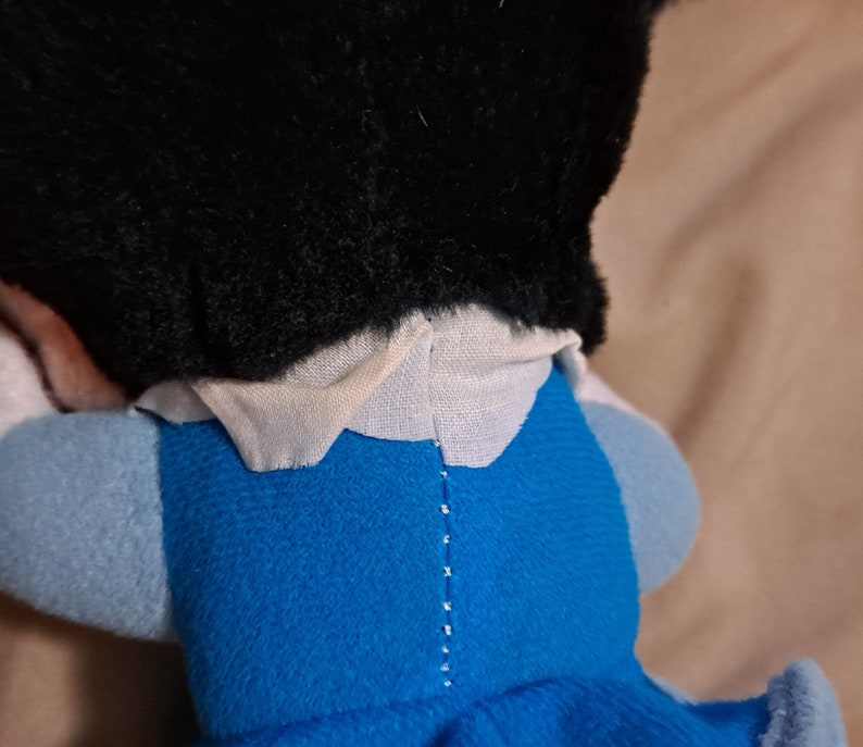 May include: A plush toy with black hair, a white collar, and a blue dress. The toy is lying on its back with its head turned to the side.