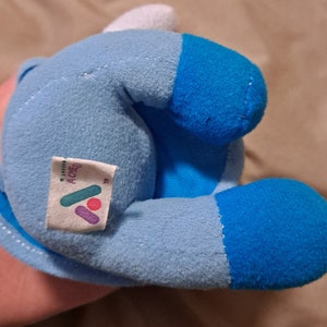 May include: A blue and white plush toy with a tag that says "ACME" on it. The toy is in the shape of a cartoon character.