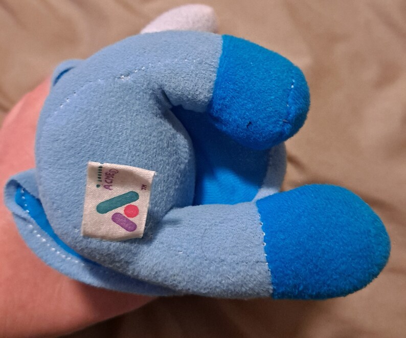 May include: A blue and white plush toy with a tag that says "ACME" on it. The toy is in the shape of a cartoon character.
