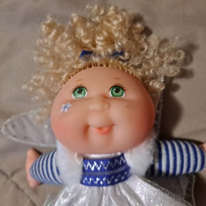 May include: A Cabbage Patch Kid doll dressed as a fairy with blonde curly hair, a white dress with blue trim, and blue and white striped sleeves. The doll has green eyes and a blue bow in her hair.