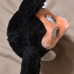 May include: A black and orange plush toy with large eyes. The toy has a black furry head and body with orange fur on the face and a white patch on the side.